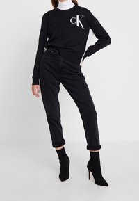 Svart stickad tröja med en vit sidpanel och "CK"-logotyp, ihop med upprullade svarta jeans och svarta ankelstövlar.