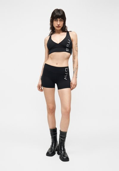 Femme aux cheveux foncés portant un soutien-gorge de sport noir et un short assorti avec un texte blanc, associée à des bottes noires mi-mollet, debout devant un fond blanc.
