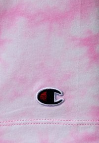 Różowa tkanina bawełniana w stylu tie-dye z małym wyszywanym logo w czarnych i czerwonych akcentach. Widoczne detale szwów wzdłuż krawędzi.