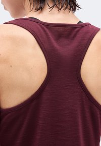 Zbliżenie na górną część pleców osoby noszącej bezrękawnik bordowego koloru typu racerback, z widocznymi krótkimi ciemnymi włosami u nasady karku.