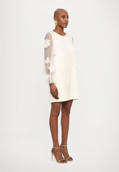 Korte witte jurk met doorschijnende, lange mouwen versierd met bloemapplicaties. Model draagt naaktkleurige hoge hakken. Eenvoudig, elegant ontwerp.