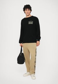 Zwarte gebreide trui met een logo, beige sweatpants met zijgraphics en witte sneakers. De persoon houdt een zwarte tas vast.