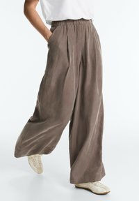 Personne portant un pantalon marron ample à jambes larges avec taille élastique et des baskets blanches, debout avec une jambe légèrement levée.