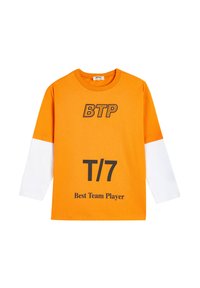 Oranssi puuvillainen pitkähihainen t-paita valkoisilla hihoilla. Siinä on mustalla painettuna "BTP", "T/7" ja "Paras tiimipelaaja". Normaali istuvuus.