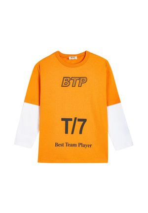 Oranssi puuvillainen pitkähihainen t-paita valkoisilla hihoilla. Siinä on mustalla painettuna "BTP", "T/7" ja "Paras tiimipelaaja". Normaali istuvuus.