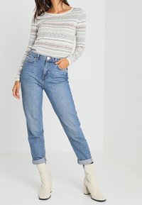 Blusa de manga larga con estampado de diseños geométricos multicolores, acompañada de unos vaqueros de cintura alta de color azul claro y botas blancas de tobillo.