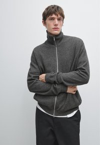 Ung man med brunt hår, iklädd en mörkgrå zip-up-tröja, vit undertröja och svarta byxor, som står mot en enkel ljus bakgrund.
