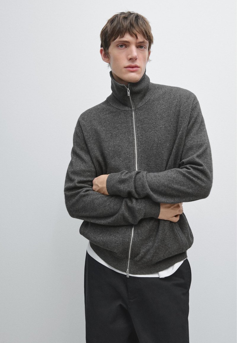 Ung man med brunt hår, iklädd en mörkgrå zip-up-tröja, vit undertröja och svarta byxor, som står mot en enkel ljus bakgrund.