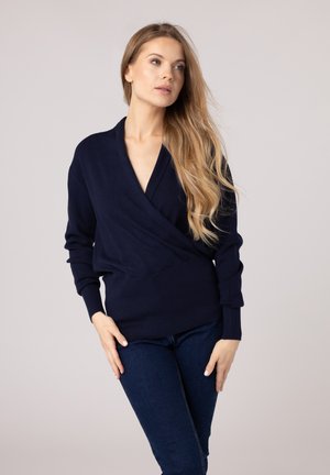 Navy wrap sweater with long sleeves and deep V-neck. Features soft knit texture and fitted waistband. Paired with dark jeans.