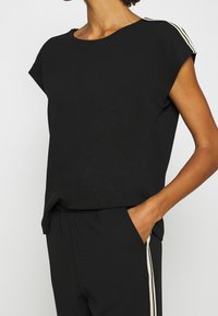 Czarny krótki top z rękawem, okrągłym dekoltem, z dwoma złotymi paskami na ramionach. Miękka, lekka tkanina; luźny krój.