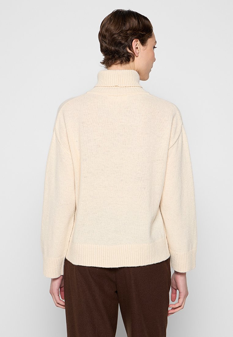 Person mit kurzen braunen Haaren trägt einen cremfarbenen Rollkragenpullover und dunkle braune Hosen, von hinten vor einem schlichten Hintergrund gezeigt.