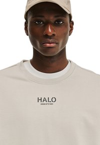 Jasno beżowy sweatshirt z okrągłym dekoltem, krótkimi rękawami i czarnym nadrukiem "HALO 198619721981" na przedzie. Gładka faktura.