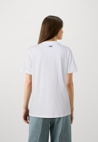 Weißes Baumwoll-T-Shirt mit kurzen Ärmeln, auf der Rückseite im Nackenbereich mit einem blauen Logo. Glatte Textur, lässige Passform. Kombiniert mit hellblauen Hosen.