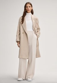 Beige wollen jas met een reverskraag, dubbele knopen, gecombineerd met een witte coltrui en crèmekleurige high-waisted broek. Witte sneakers.