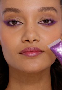 Paarse cosmetische gel in een heldere tube tegen een gezicht gehouden met paarse oogmake-up en glanzende roze lippen, met een soepele huid en een natuurlijke textuur.