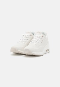 Hvite sneakers med en teksturert overdel, som har ventilasjonsåpninger, runde snørebånd og en synlig luftdempningsenhet i sålen.