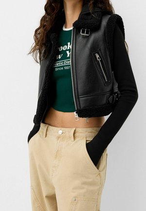Persoon draagt een zwart shearlingvest over een groen cropped shirt en beige hoog getailleerde broek met handen in de zakken.