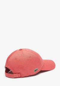 Gorra de algodón color coral con visera curva y correa ajustable. Cuenta con un logo de cocodrilo bordado en verde en el lateral. Textura suave.