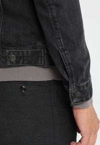 Chaqueta de mezclilla negra con puños y cinturilla acanalados en gris. Presenta un cierre de botones y un acabado texturizado en la tela.