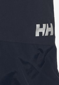 Mörkblå plagg med texturerad nederdel, med en glänsande silverfärgad "HH"-logotyp på övre höger sida. Materialet verkar hållbart och vattenavvisande.