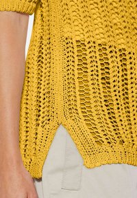 Gelber gestrickter Pullover mit offener Masche, featuring einen weiten Ausschnitt und Seitenschlitze. Rippbündchen und Saum verleihen dem Design Textur.