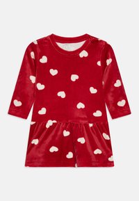 Robe en velours rouge avec des manches longues, motifs de cœurs blancs et jupe froncée. Elle comporte un col rond et une fermeture à bouton à l'épaule.