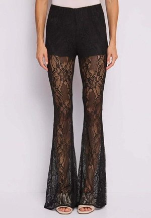 Pantaloni neri in pizzo a vita alta con motivo floreale, gambe svasate semi-trasparenti, indossati con sandali beige open-toe.