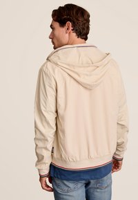 Veste à capuche beige clair avec un ourlet côtelé, ornée d'accents rayés rouges et bleus et d'une texture lisse. Visible de dos.