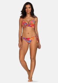 Kobieta o ciemnych włosach, ubrana w kolorowy kwiecisty bikini z wiązaniami po bokach, stoi boso na jasnoszarym tle.