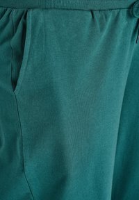 Mėlynos jogger tipo kelnės, pagamintos iš minkštos medvilnės medžiagos, su elastinga juosmeniu ir šoninėmis kišenėmis, pasižyminčios lygiu, tolygiai išsiskiriančiu tekstūros paviršiumi.