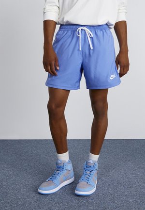 Persoon draagt blauwe Nike short met wit trekkoord, witte sokken en blauwgrijze Nike hoge sneakers, staand op een grijs tapijt.