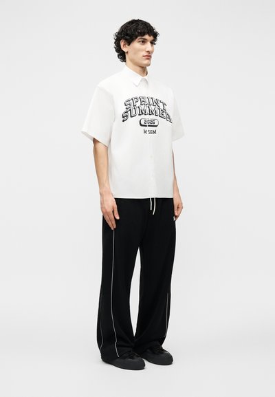 Jeune homme aux cheveux bouclés portant une chemise blanche à manches courtes avec le texte "SPRINT SUMMER 2026" et un pantalon large noir avec un liseré blanc.