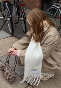 Witte wollen sjaal met franjes, gelabeld "ARCTIC FOX", gedragen bij een beige jas en vastgehouden naast een gestructureerde, patroonhandtas. Twee fietsen op de achtergrond.