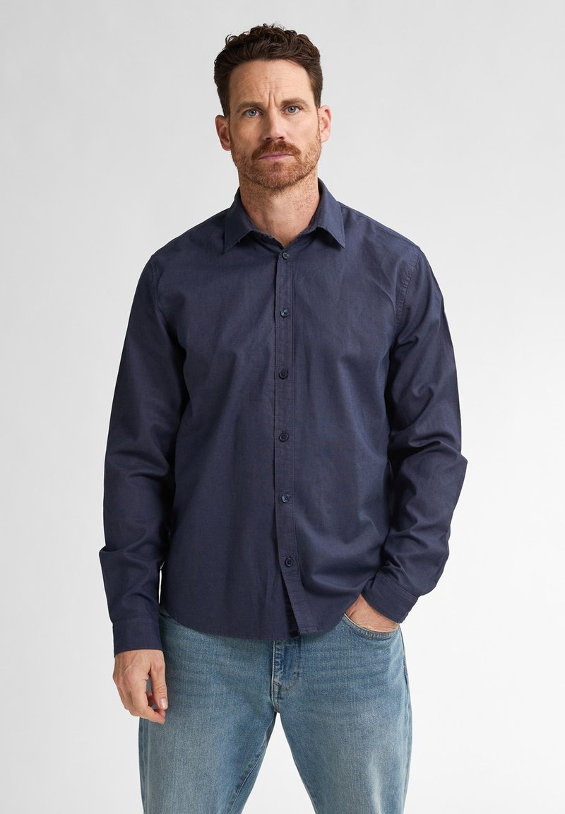 Miestens tamsiai mėlyna marškinė su mygtukais, pagaminta iš lengvos medžiagos. Turi ilgomis rankovėmis, su mygtukais ant riešo ir klasikiniu apykaklės dizainu. Dėvimi su šviesiomis džinsinėmis kelnėmis.