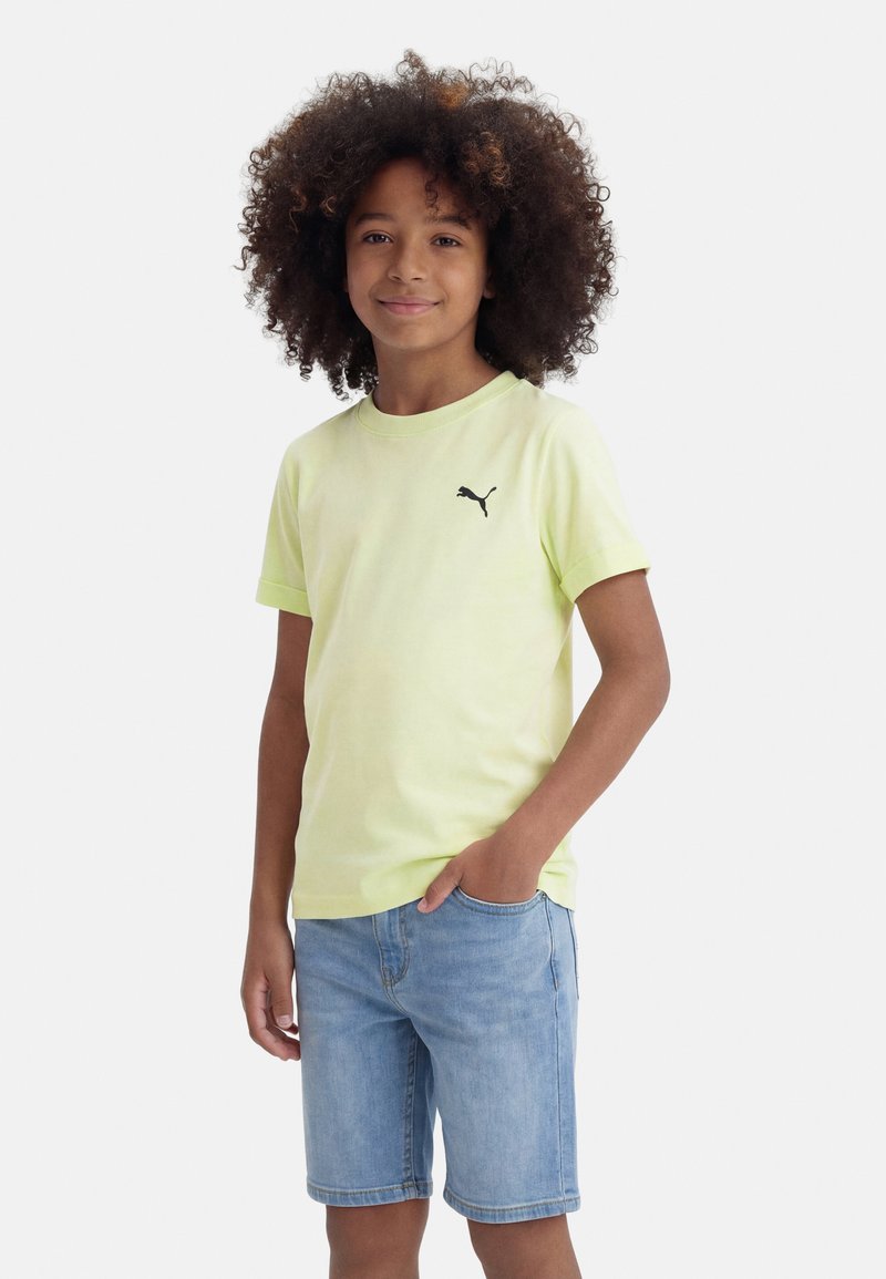 Παιδί με κατσαρά μαλλιά, φοράει ανοιχτό κίτρινο μπλουζάκι Puma και τζιν σορτς, στέκεται με το ένα χέρι στην τσέπη, χαμογελώντας ελαφρώς.