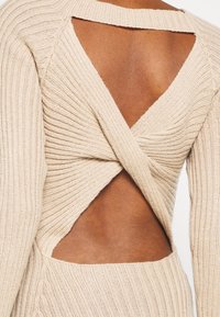 Pull beige en tricot avec un design croisé dans le dos. Caractéristiques : texture côtelée, sections ouvertes et silhouette ajustée.