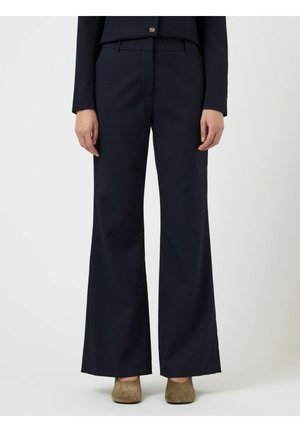 Kvinde iført marineblå vidde bukser og brune ruskindssko, stående mod en enkel lyse baggrund.