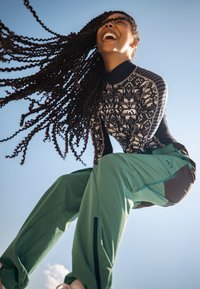 Haut à manches longues noir et blanc à motif avec un col zippé, associé à un pantalon ample vert et marron. Fond de ciel bleu clair.