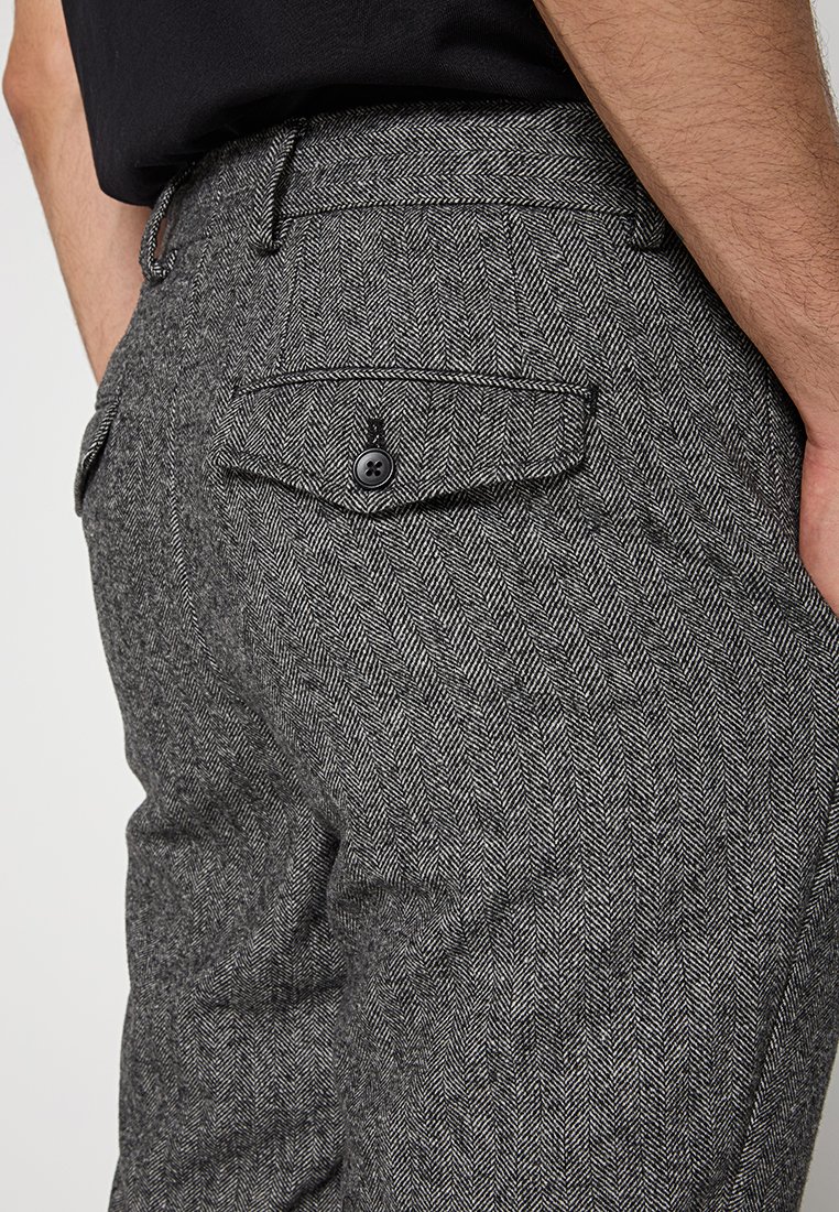 Ripresa ravvicinata di una persona che indossa pantaloni grigi a spina di pesce con una tasca posteriore con bottone e una camicia nera infilata dentro.