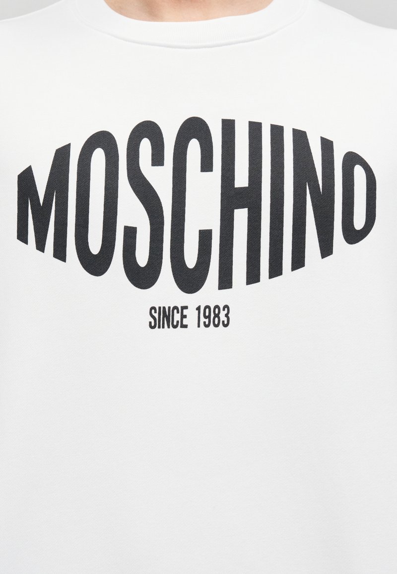 Sweatshirt en coton blanc avec le texte noir audacieux "MOSCHINO" et "SINCE 1983" imprimé en dessous. Col rond et coupe décontractée.