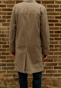 Trench coat beige con taglio dritto, apertura singola sul retro e polsini abbottonati, indossato sopra un denim scuro. Tessuto testurizzato.