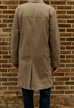 Trench coat beige con taglio dritto, apertura singola sul retro e polsini abbottonati, indossato sopra un denim scuro. Tessuto testurizzato.