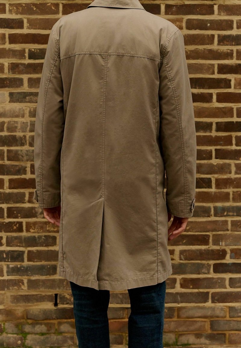 Trench coat beige con taglio dritto, apertura singola sul retro e polsini abbottonati, indossato sopra un denim scuro. Tessuto testurizzato.