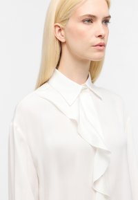 Blusa di seta bianca con colletto strutturato e dettagli di volant a cascata lungo il davanti, dotata di maniche lunghe e una texture liscia.