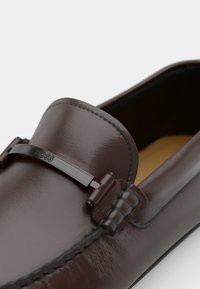 Mocassins en cuir marron avec une finition texturée, un accent logo en métal noir et des détails cousus sur le bord supérieur.