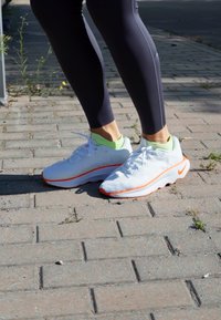 Fehér sportcipők narancs szegéllyel, hálós felsőrész és párnázott talp jellemzi őket. Fekete leggings-szel viselve, élénk zöld belső gallérral.