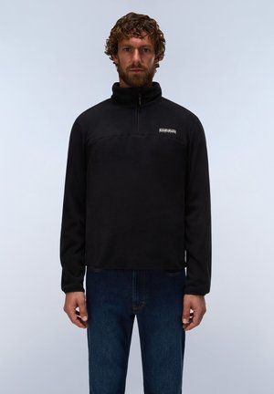 Man with curly hair and beard wearing black zip-up fleece jacket and blue jeans, standing against plain light background.