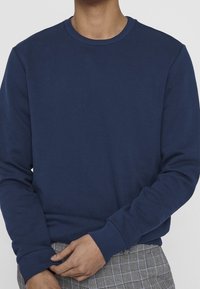 Granatowy sweter z długimi rękawami, z prążkowanym dekoltem i mankietami, wykonany z miękkiego materiału, w zestawie z szarymi spodniami w kratę.