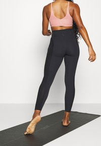 Czarne legginsy z wysokim stanem z teksturowanym panelem bocznym, połączone z różowym stanikiem sportowym. Osoba stoi na czarnej macie do jogi.