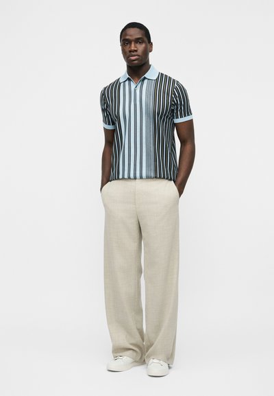 Άνδρας στέκεται με τα χέρια στις τσέπες, φοράει κοντομάνικο μπλε και μαύρο πόλο με κάθετες ρίγες και μπεζ παντελόνι με φαρδιά μπατζάκια, και λευκά παπούτσια.